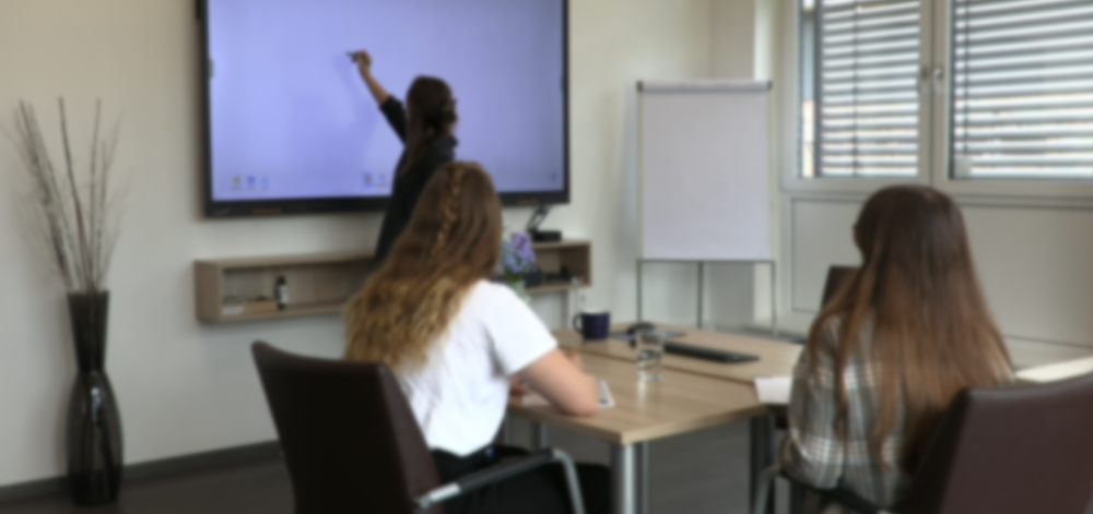 Zwei Mitarbeiterinnen sitzen im Meetingraum und eine weitere schreibt etwas auf das Whiteboard